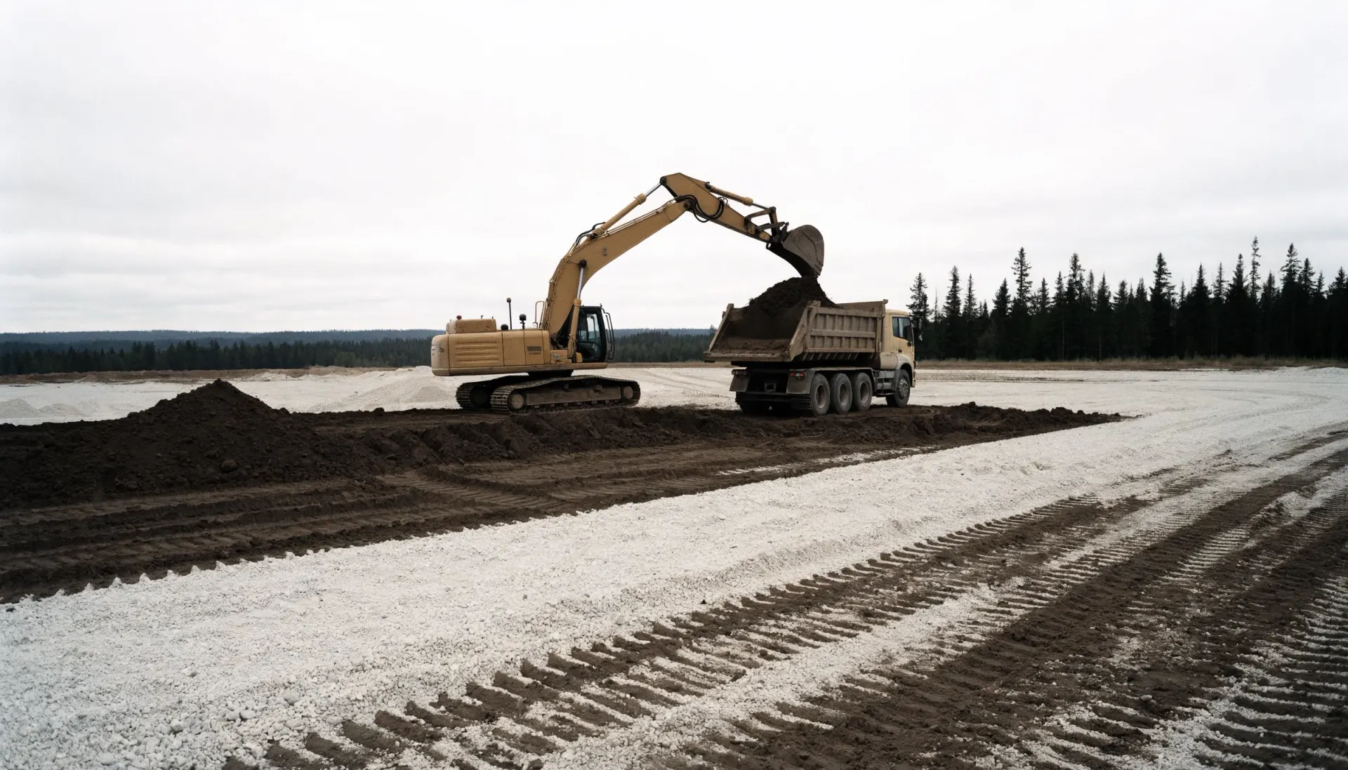 Excavatrice chargeant de la terre dans la benne d’un camion sur un chantier préparé, sous un ciel couvert, lisière d’épinettes à l’horizon.