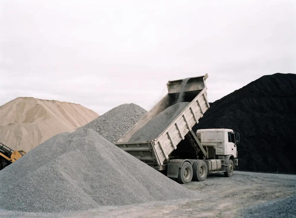 Camion à benne déversant une charge de gravier gris en cône dans une cour de matériaux, piles de sable et de terre à l’arrière.
